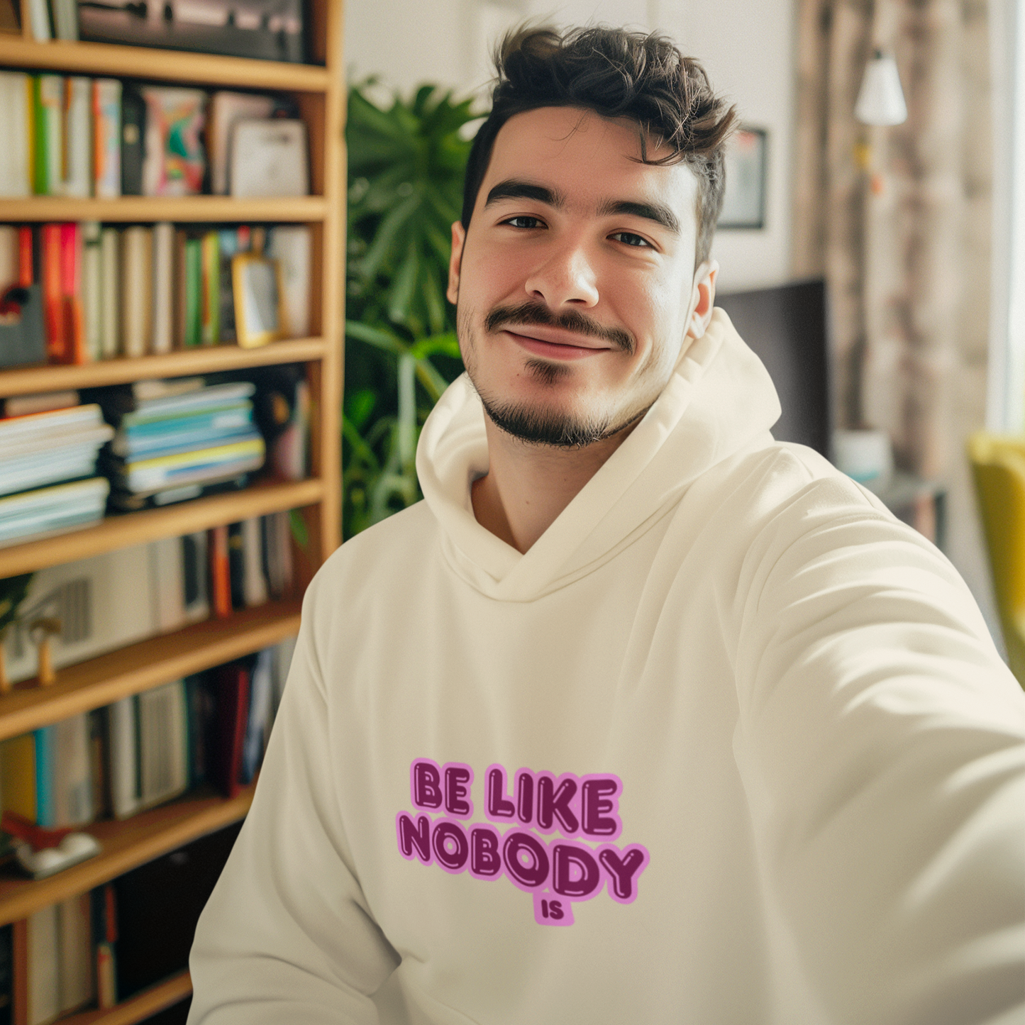 Man wearing a white hoodie with 'Be Like Nobody Else' text in a room with bookshelves and plants.