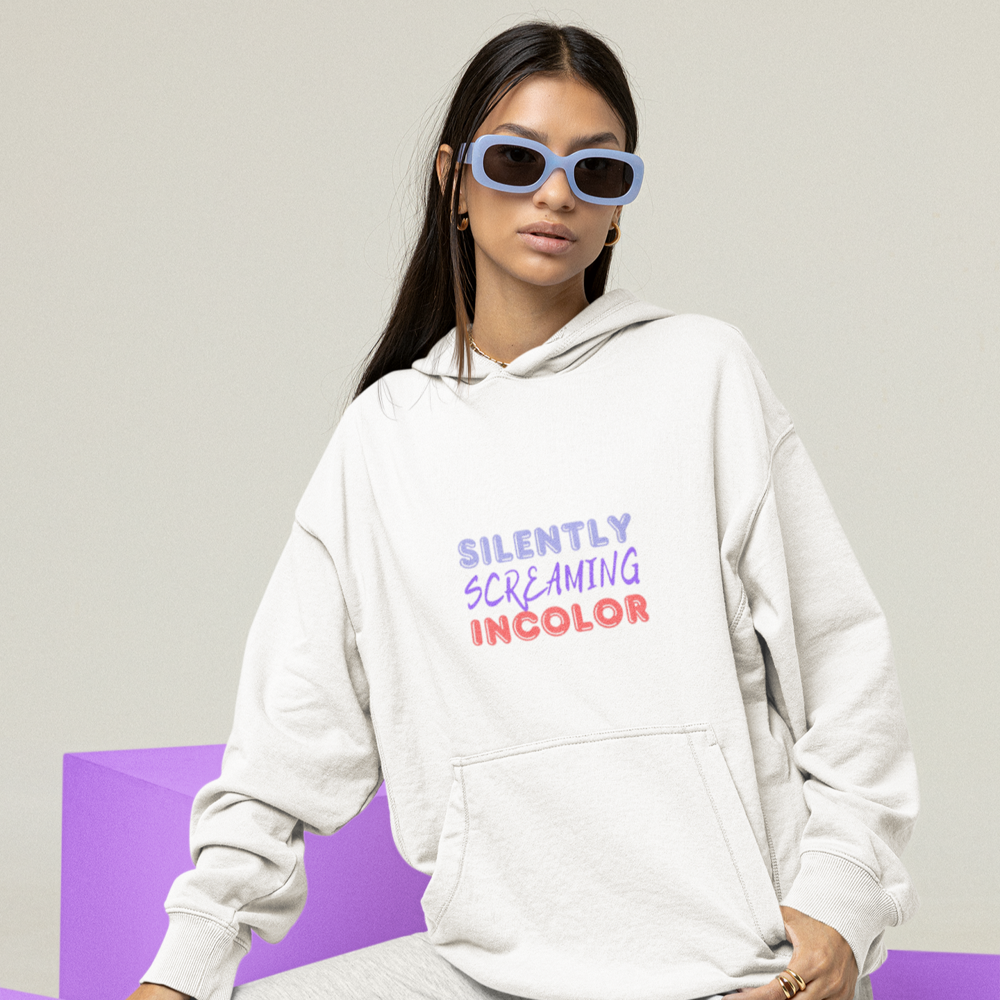 Person wearing a white hoodie with colorful text, sitting on a purple block against a light gray background