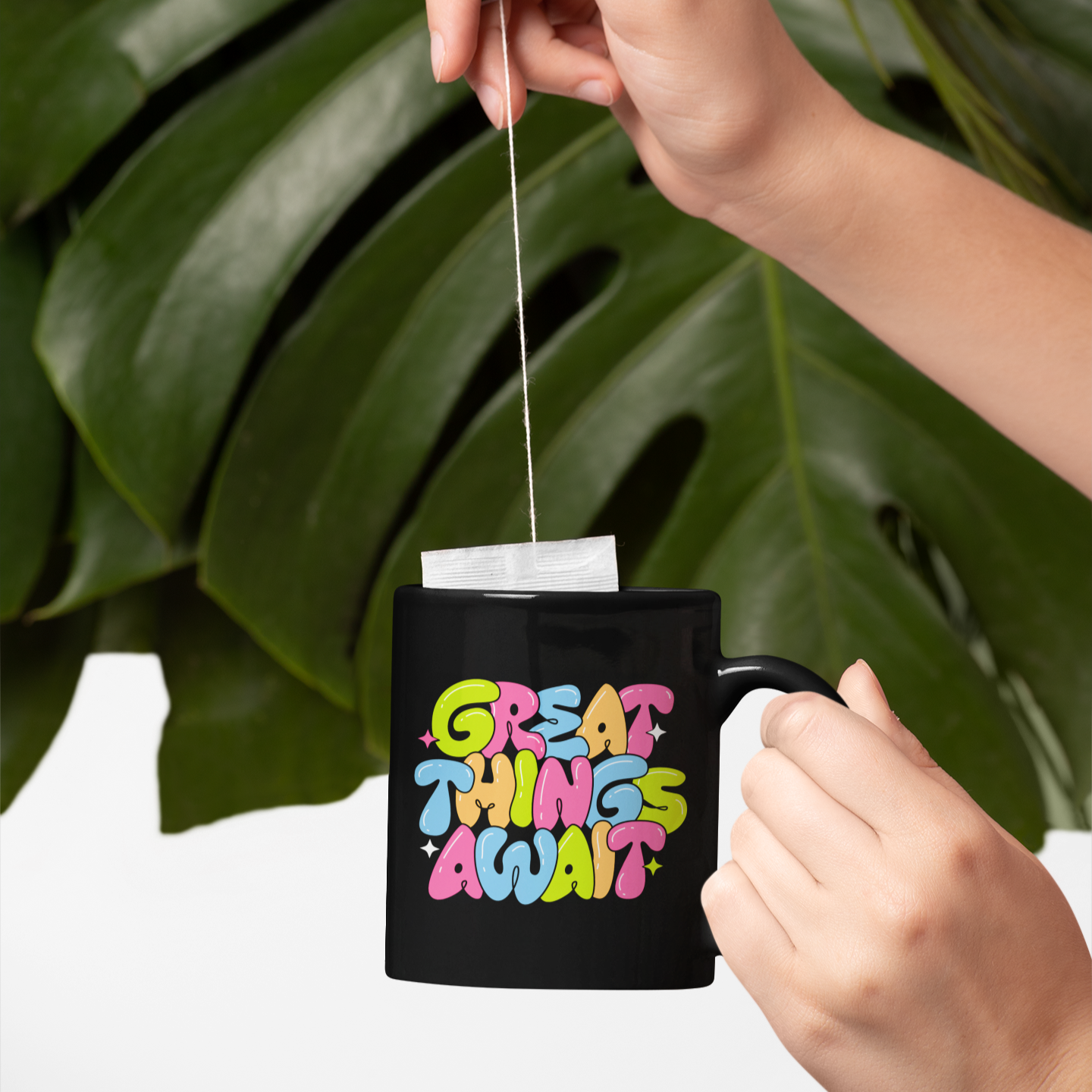 Person placing a tea bag into a black mug with 'Great Things Await' text, against a leafy background.