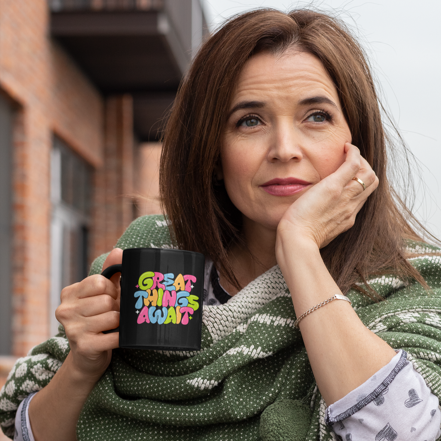 Woman holding a mug with 'Great Things AwaT' holding a green patterned scarf, standing in an urban setting.