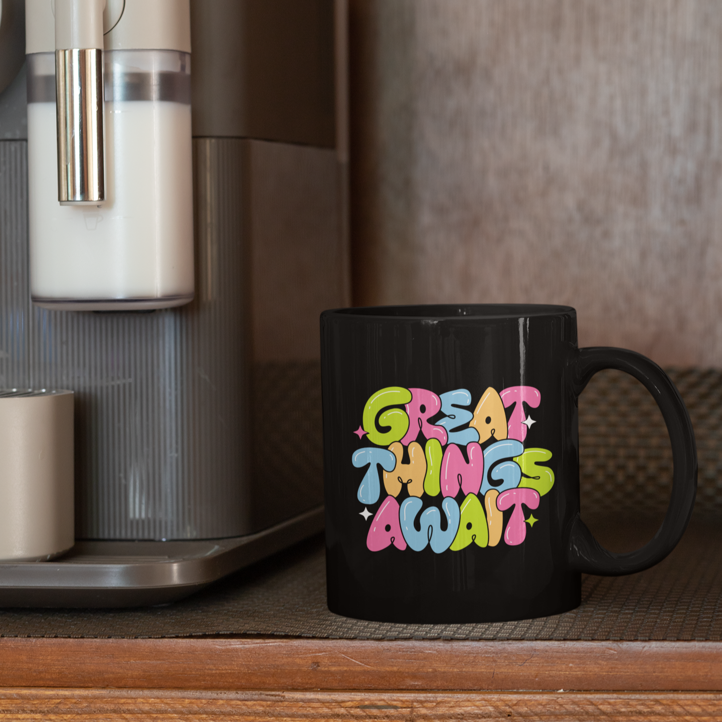 Black mug with colorful text 'Great Things Await' next to a coffee maker.