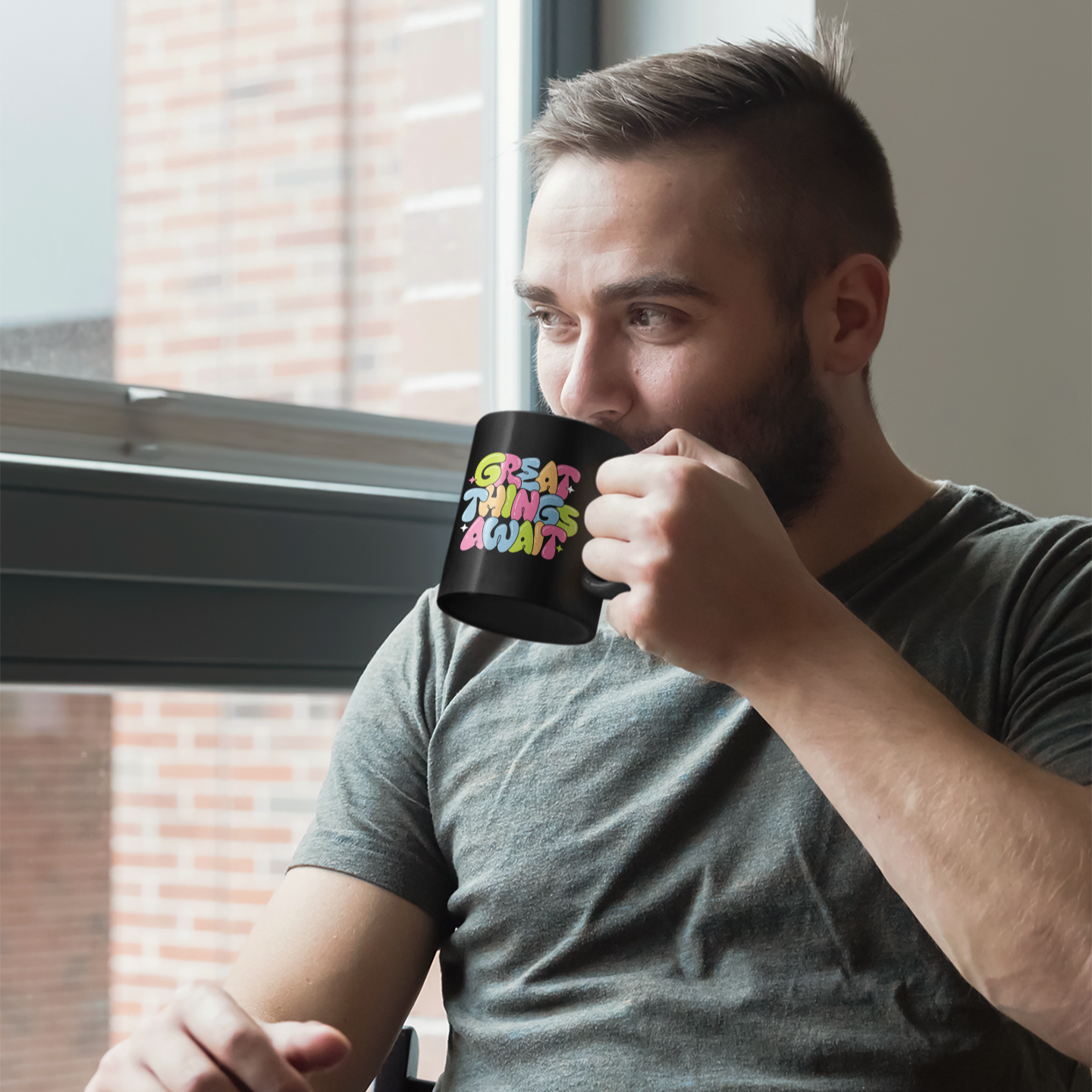 Man holding a black mug with text, sitting by a window.
