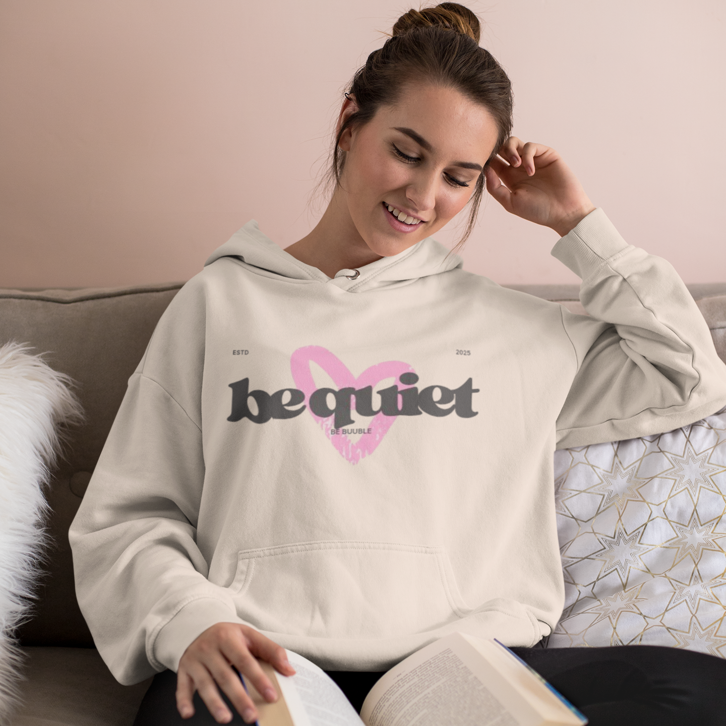 Woman wearing a hoodie with 'be quiet' text, sitting on a couch with an open book.