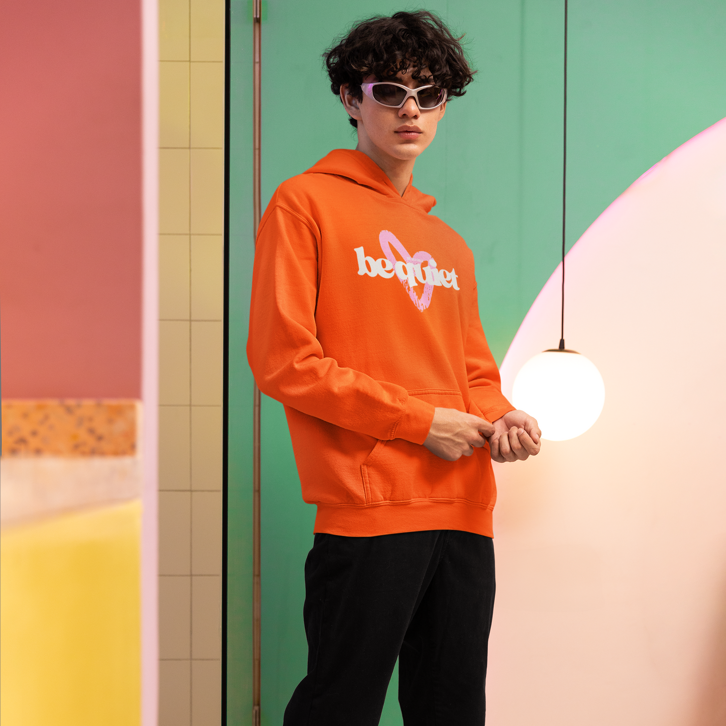 Person wearing an orange hoodie with a logo, black pants, and white sneakers in a colorful indoor setting.