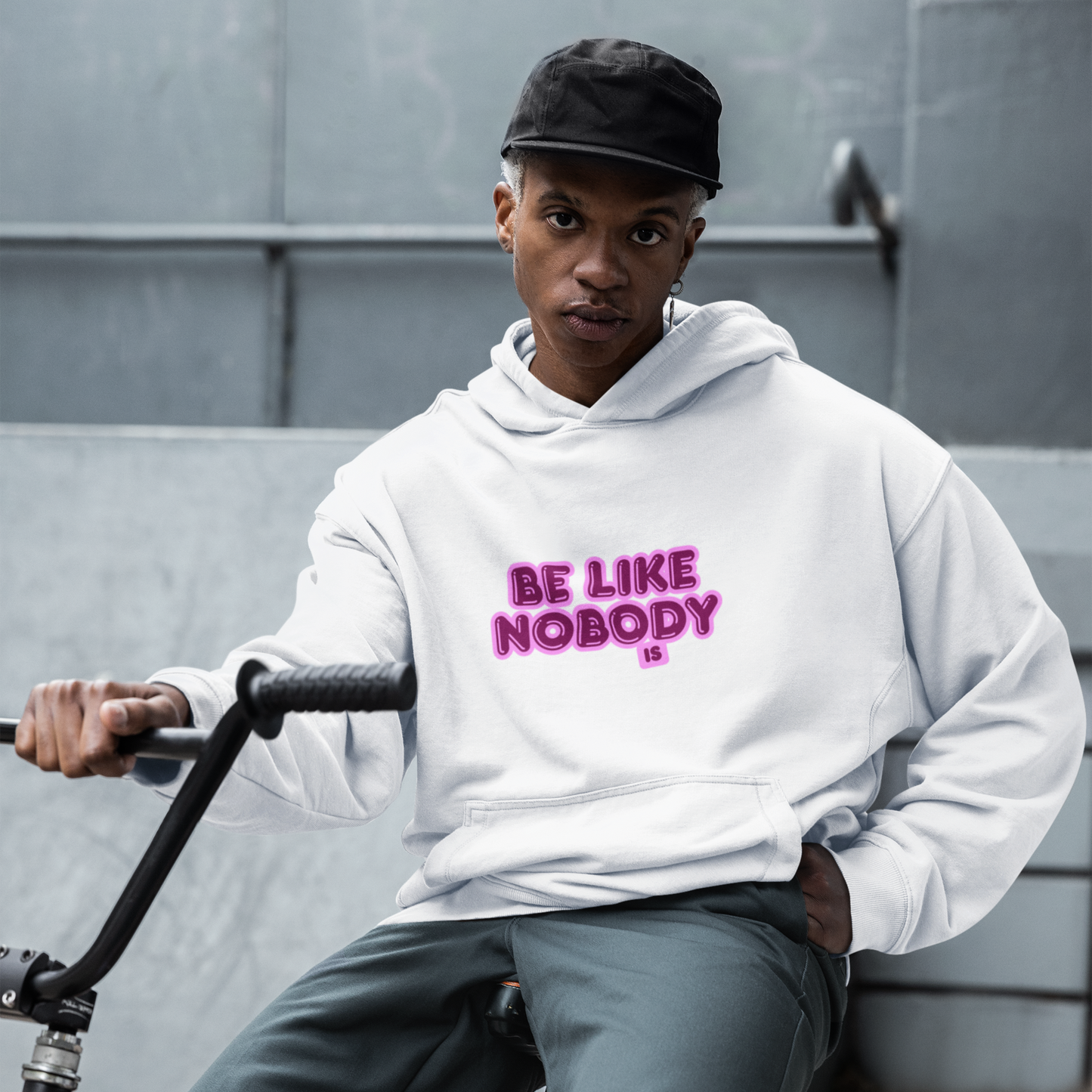 Person wearing a white hoodie with 'Be Like Nobody' text, sitting on a bike against a gray wall.