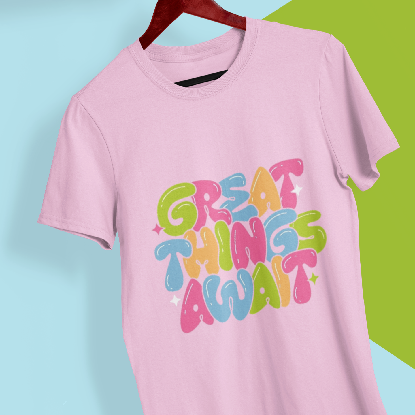 Pink t-shirt with colorful text 'Great Things Await' on a hanger against a blue and green background
