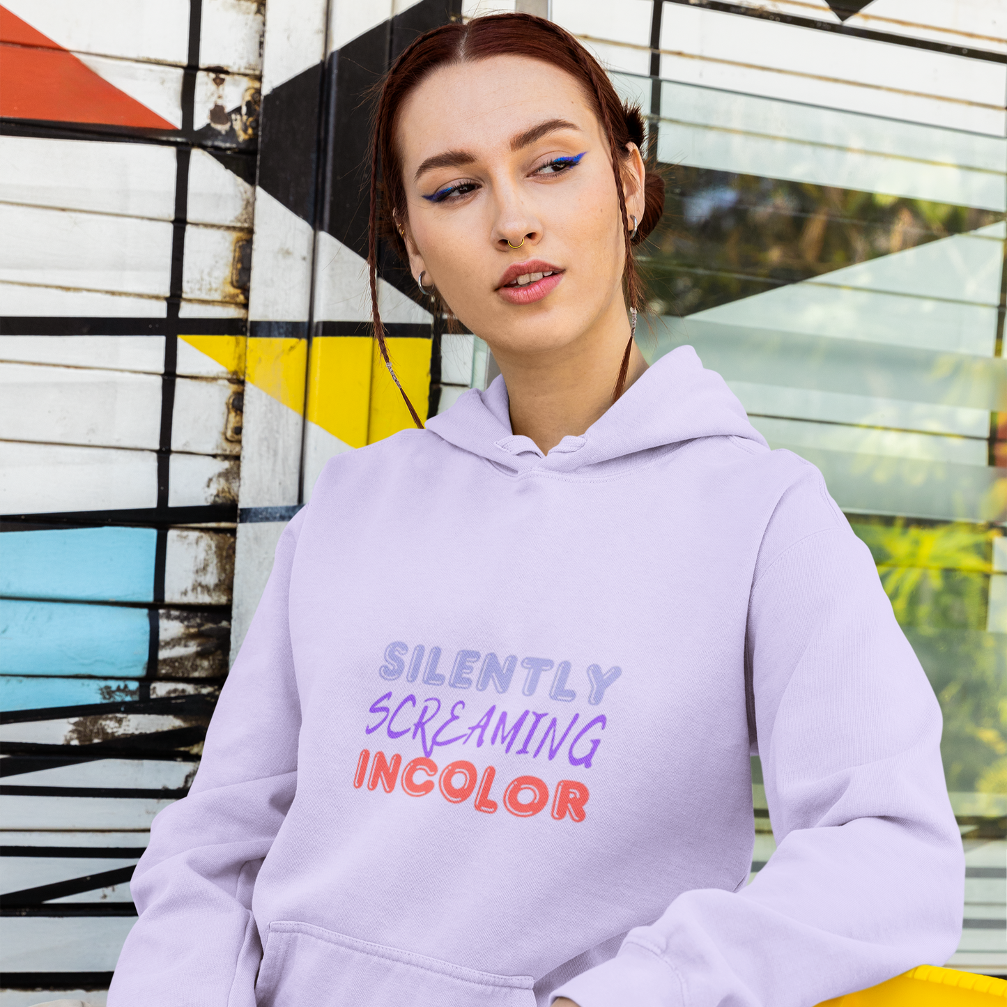Person wearing a light purple hoodie with colorful text, sitting against a geometric wall.