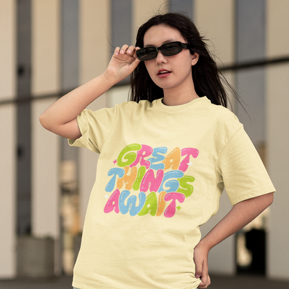 Woman wearing a light yellow t-shirt with 'Great Things Await' text, standing outdoors.