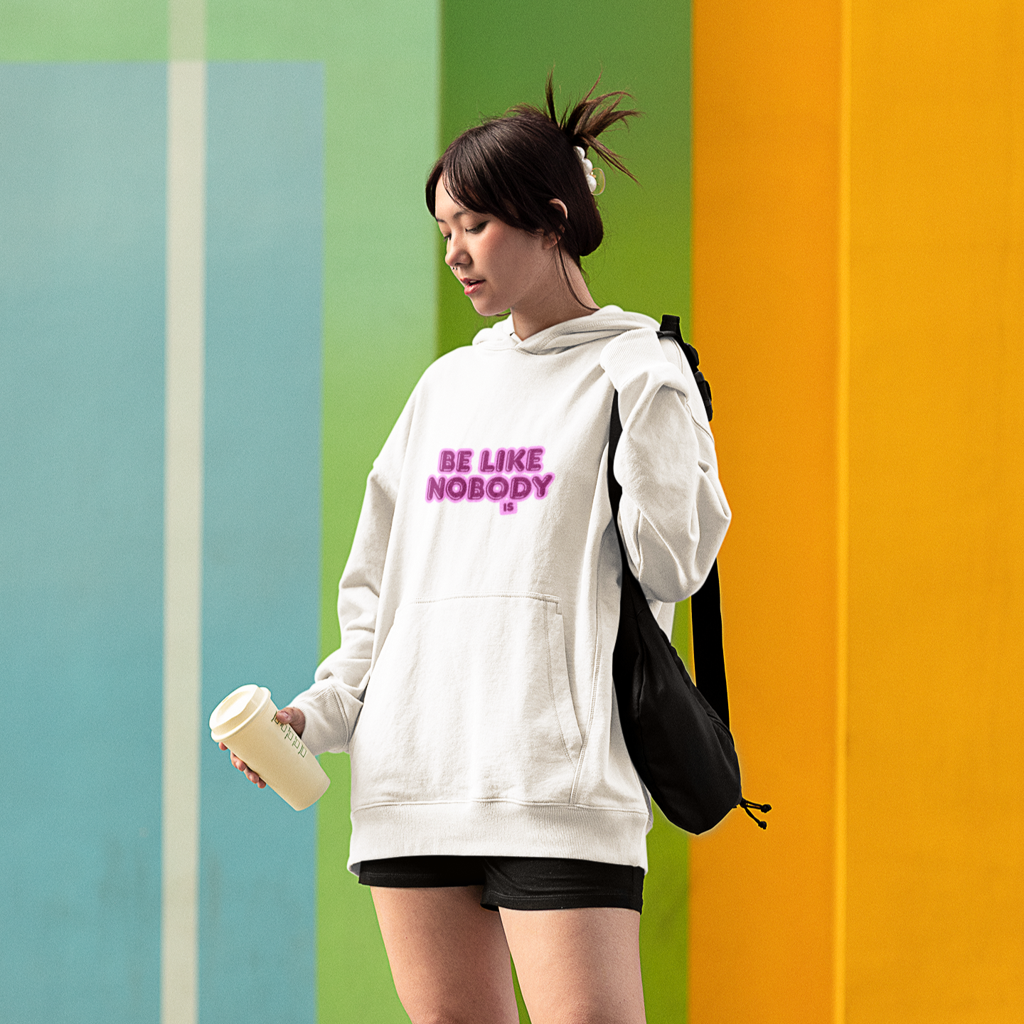 Person wearing a white hoodie with text, holding a cup and a black bag against a colorful wall.