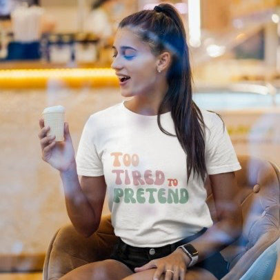 Woman sitting in a cafe holding a coffee cup, wearing a 'Too Tired to Pretend' t-shirt.