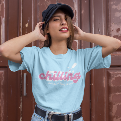 Person wearing a light blue t-shirt with 'chilling' text, denim shorts, and a cap against a brown background.
