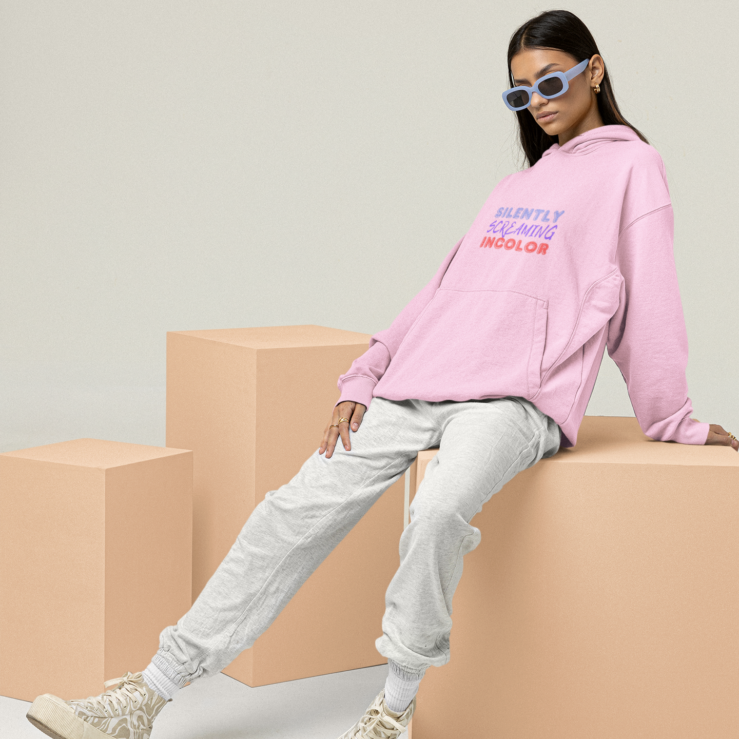 Person wearing a pink hoodie with text, sitting on a beige block against a light gray background